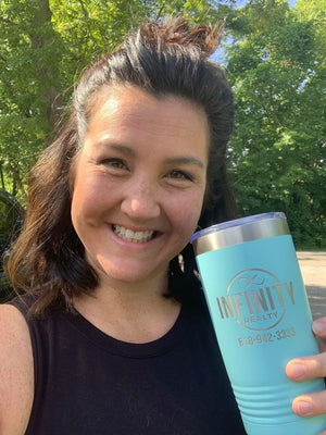 A woman with dark hair and a black tank top smiles and holds a teal tumbler with the text "INFINITY REALTY" and a phone number on it. She is outdoors with trees in the background, showcasing one of their Kodiak Coolers Custom Tumblers 20 oz with your Logo or Design Engraved - Special Bulk Wholesale Pricing - Pack of 12 Pieces - 1 Color.