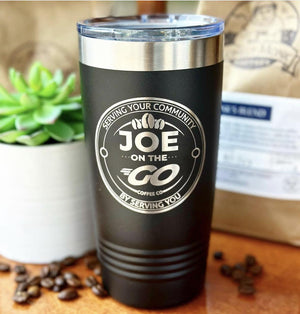 A black travel mug with the text "Joe on the Go Coffee Co - Serving Your Community by Serving You" is placed next to a potted succulent and scattered coffee beans. A bag of coffee beans is blurred in the background, emphasizing the quality of their Kodiak Coolers Custom Tumblers 20 oz with your Logo or Design Engraved - Special Bulk Wholesale Pricing - Pack of 12 Pieces - 1 Color.