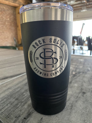A black and silver tumbler with the Rock Solid Brewing Company logo, likely one of their Kodiak Coolers Custom Tumblers 20 oz with your Logo or Design Engraved - Special Bulk Wholesale Pricing - Pack of 72 Pieces - 1 Color - $13.87 Each, is placed on a marble table inside a cozy café or bar setting.
