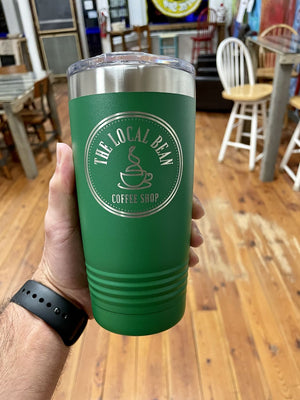 A hand holds a green travel tumbler with "The Local Bean Coffee Shop" logo in an indoor café setting with wooden floor and various chairs, showcasing Kodiak Coolers Custom Tumblers 20 oz with your Logo or Design Engraved - Special Bulk Wholesale Pricing - Bundle Packs.