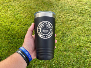 A hand holding a black, insulated tumbler with "Custom Tumblers 20 oz with your Logo or Design Engraved - Special Bulk Wholesale Pricing - Pack of 12 Pieces - 1 Color" and "Kodiak Coolers" laser engraved on it. The person is wearing blue and green wristbands. The background is grass.