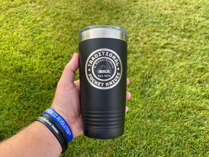 A hand holds a black tumbler with the logo "Traditional Pocket Knives" established in 1975, over a background of green grass. The person is wearing blue and black wristbands. The Kodiak Coolers Custom Tumblers 20 oz with your Logo or Design Engraved - Special Bulk Wholesale Pricing - Pack of 96 Pieces - 1 Color - $12.49 Each feature intricate details that make them truly unique.