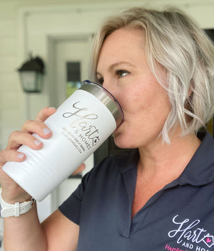 A person with short blonde hair drinks from a white, Kodiak Coolers Custom Tumblers 20 oz with your Logo or Design Engraved - Special Bulk Wholesale Pricing - Pack of 24 Pieces - 1 Color - $16.63 Each. They are wearing a navy polo shirt with a logo on the chest and standing in front of a house with a lantern on the wall.