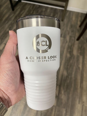 A person holding a white insulated tumbler with the text "A Closer Look Home Inspection" and a logo featuring the letters "ACL." The background shows a wooden floor and part of a chair leg. These Custom Tumblers 20 oz with your Logo or Design Engraved - Special Bulk Wholesale Pricing - Bundle Packs from Kodiak Coolers are perfect for promoting your business in style.