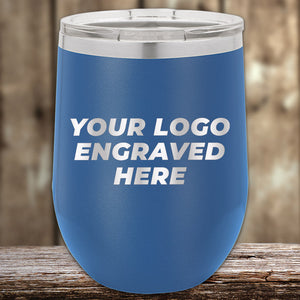 Positioned on a wooden surface with a blurred wood background, the Kodiak Coolers' blue custom tumbler features laser-etched "Your Logo Engraved Here" text. Perfect as a corporate promotional gift, it’s offered with no setup fees for a limited time.