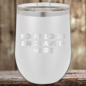 A white Kodiak Coolers custom tumbler with a lid, engraved with "YOUR LOGO HERE," sits on a wooden surface. It's perfect for corporate gifts, blending style with personalization. Don't miss this limited-time offer on our laser-etched design with no setup fees!.