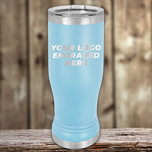 A blue Custom Pilsner Tumbler 14 oz from Kodiak Coolers stands upright on a wooden surface with the text "YOUR LOGO ENGRAVED HERE" written on it. The background is blurred, showing wooden planks. Perfect for bulk wholesale pricing, it offers an excellent way to showcase your brand with an engraved logo.