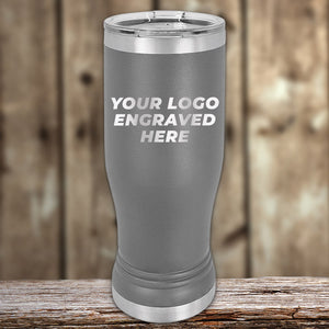 A grey stainless steel tumbler with a metal lid, featuring the text "YOUR LOGO ENGRAVED HERE" on its side. The background is a wooden surface and a rustic wood panel backdrop. Ideal for companies seeking bulk wholesale pricing or Custom Pilsner Tumblers 14 oz with your Logo or Design Engraved | No Minimal Order | Sample Volume Pricing from Kodiak Coolers.