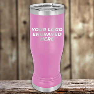 A pink stainless steel tumbler with silver accents stands on a wooden surface, featuring the text "Your Logo Engraved Here." Ideal for personalized branding, it offers a sleek alternative to Custom Pilsner Tumblers 14 oz with your Logo or Design Engraved by Kodiak Coolers.