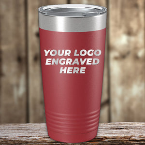 A red Kodiak Coolers Custom Tumblers 20 oz with your Logo or Design Engraved with a silver lid and the words "YOUR LOGO ENGRAVED HERE" written on it, showcasing its laser engraved details, is placed on a wooden surface with a blurred wooden background. Perfect for those seeking custom tumblers at wholesale pricing.