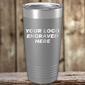 A silver tumbler with the text "YOUR LOGO ENGRAVED HERE" against a wooden background, perfect for wholesale custom tumblers or bulk orders. Introducing the Custom Tumblers 20 oz with your Logo or Design Engraved - Special Bulk Wholesale Pricing - Pack of 96 Pieces - 1 Color - $13.53 Each by Kodiak Coolers.