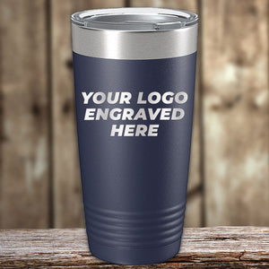 A dark blue travel tumbler with a silver lid displayed on a wooden surface. The text on the tumbler reads "YOUR LOGO ENGRAVED HERE." Perfect for those seeking custom tumblers, each Kodiak Coolers Custom Tumbler 20 oz with your Logo or Design Engraved - Special Bulk Wholesale Pricing - Bundle Packs is laser engraved to ensure a lasting imprint.