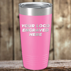 A pink travel tumbler with a silver lid sits on a wooden surface. The front of the tumbler says, "YOUR LOGO ENGRAVED HERE" in white text. This Custom Tumblers 20 oz with your Logo or Design Engraved - Special Bulk Wholesale Pricing - Bundle Packs by Kodiak Coolers can be laser engraved and is available at wholesale pricing. The background features blurred wooden paneling.