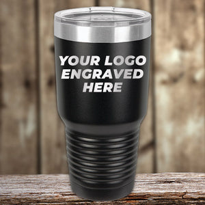 A black travel tumbler with the text "YOUR LOGO ENGRAVED HERE" displayed on it, set against a rustic wooden background—a perfect example of Kodiak Coolers' Custom Tumblers 30 oz with your Logo or Design Engraved | No Minimal Order | Sample Volume Pricing, ideal for promotional items.