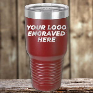 A red Kodiak Coolers Custom Tumbler 30 oz with your Logo or Design Engraved and a clear lid sits on a wooden surface, featuring the text "YOUR LOGO ENGRAVED HERE" on its front. These custom tumblers make perfect promotional items for your brand.