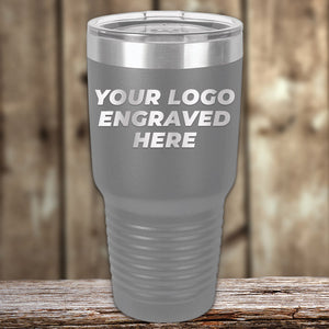 A grey Custom Tumblers 30 oz with your Logo or Design Engraved | No Minimal Order | Sample Volume Pricing from Kodiak Coolers with a clear lid sits on a wooden surface. The custom tumbler features the text "YOUR LOGO ENGRAVED HERE" on its front. The background is a blurred wooden wall, making it perfect for promotional items.