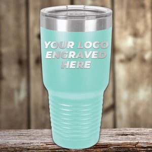 A light blue travel tumbler with a silver lid and the text "Kodiak Coolers Custom Tumblers 30 oz with your Logo or Design Engraved | No Minimal Order | Sample Volume Pricing" embossed on the front, positioned on a wooden surface. These custom tumblers make excellent promotional items for your brand.