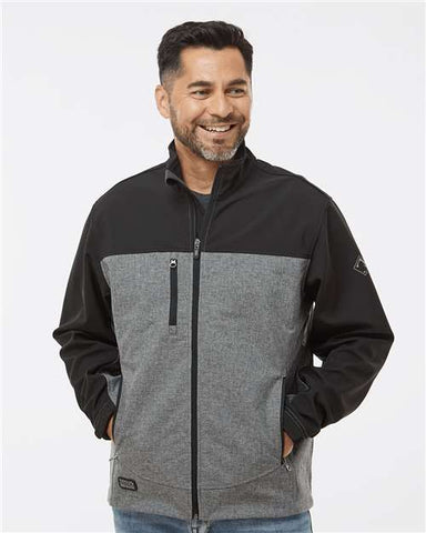 A man wearing a black and gray Motion DRI DUCK Soft Shell Jacket with his hands in his pockets, smiling, against a plain white background. This stylish, wind-and-water-resistant jacket from DRI DUCK is available with customized options and wholesale pricing.