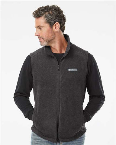 A man in a Columbia Steens Mountain Fleece Vest by Columbia, dark gray, layered over a black long-sleeve shirt, is looking to his left with his hands tucked into the vest pockets—an ideal ensemble for outdoor activities.