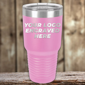 A pink insulated tumbler from Kodiak Coolers, featuring a silver lid, rests on a wooden surface against a rustic backdrop. The front is boldly engraved with "Your Logo Engraved Here" in silver letters. Ideal for custom mugs or promotional gifts, this drinkware combines style and functionality. Don't miss the SPECIAL 72 HOUR SALE PRICING that includes single-side engraving!